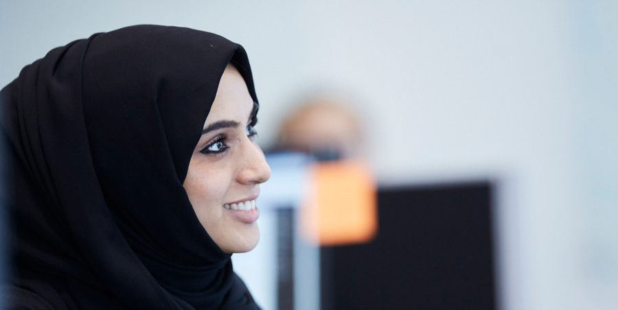 With a pleasant expression, a woman wearing a hijab smiles and directs her gaze away from the camera. The photograph is a side angel of her face.
