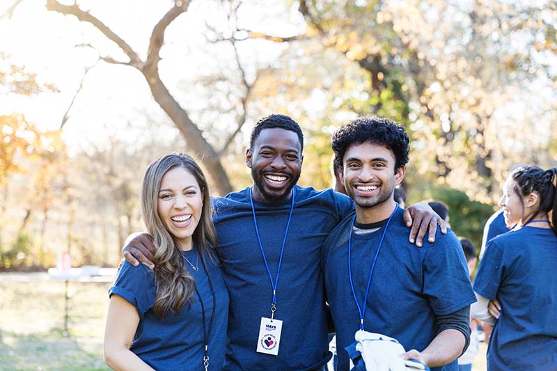 Three individuals - two male and one female - wear the same blue t-shirt and lanyards around their necks. They smile brightly for a group photo.  It's an outdoor setting with trees in the background.