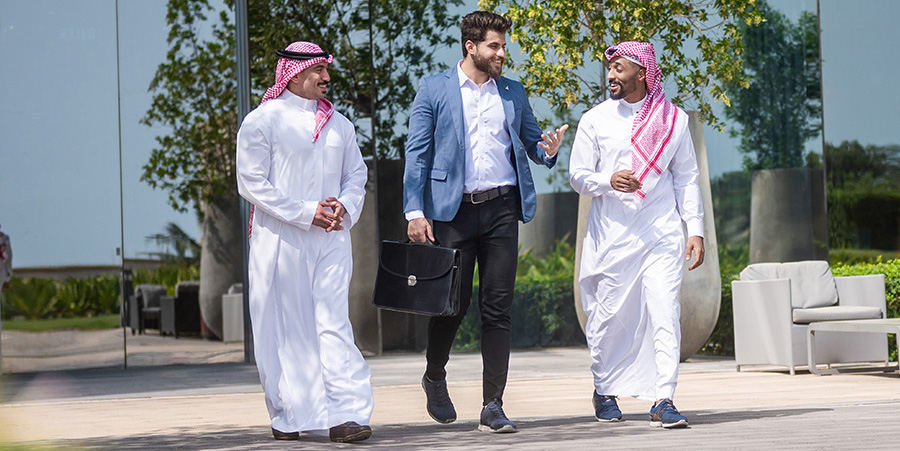 Three men walking outside a modern building. Two of the men wear traditional Middle Eastern attire and walk beside a man in business attire carrying a briefcase and wearing a blue blazer.
