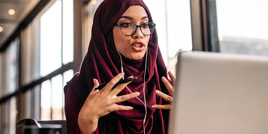 A woman wearing reading glasses and a purple hijab looks animated as she speaks while looking at a laptop or desktop screen, potentially in an online meeting.
