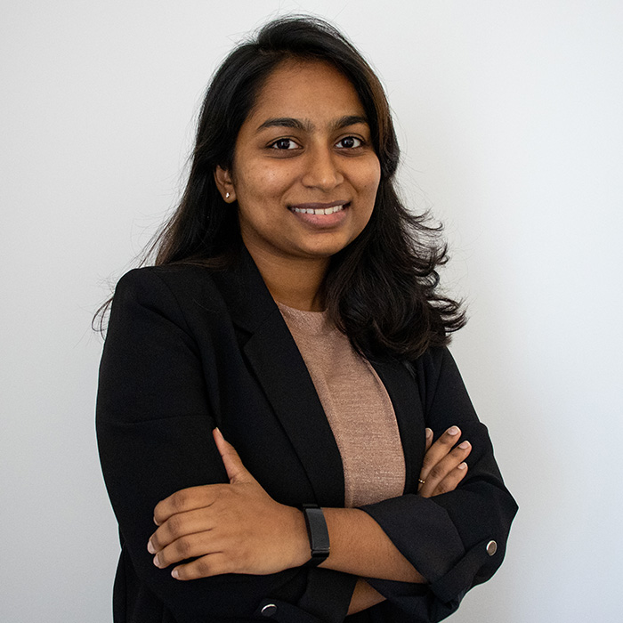 A woman with dark hair is standing against a plain white background. She is wearing a black blazer, a brown top, and a black wristwatch, with arms crossed, smiling.