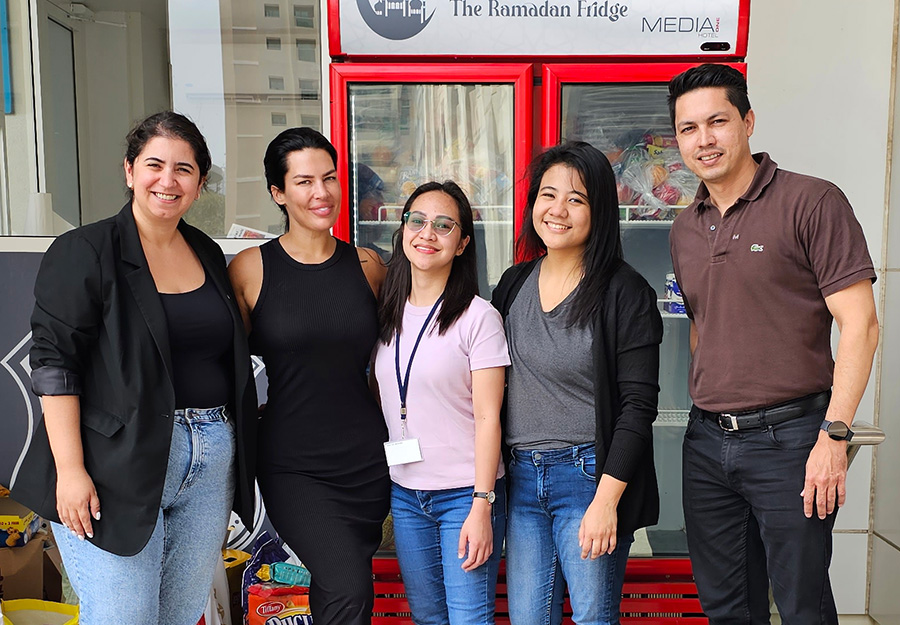 A group of individuals partake in a charity initiative. They stand in front of a Ramadan Fridge for a food drive. 