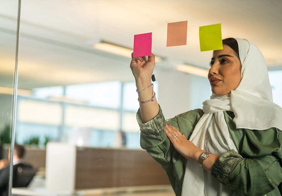 A woman wearing a hijab writes on a post-it note. She appears focused and determined.