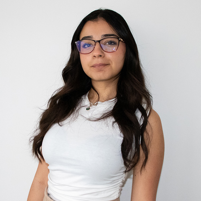 A young woman wearing glasses, dressed in a white shirt is photographed smiling softly and looking directly at the camera. Her hair is slightly longer than shoulder length. She wears a gold locket. The photograph is a headshot.