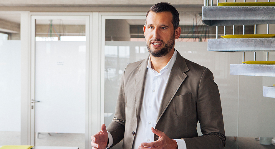 A smartly dressed man is photographed candidly while speaking and kindly guesturing with his hands. He wears a beige blazer and a white shirt unbottoned at the collar.