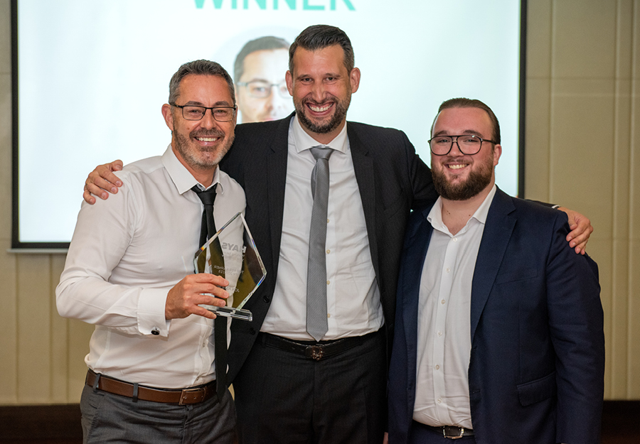 Three formally dressed gentlemen proudly display an award, smiling broadly for a group photo.