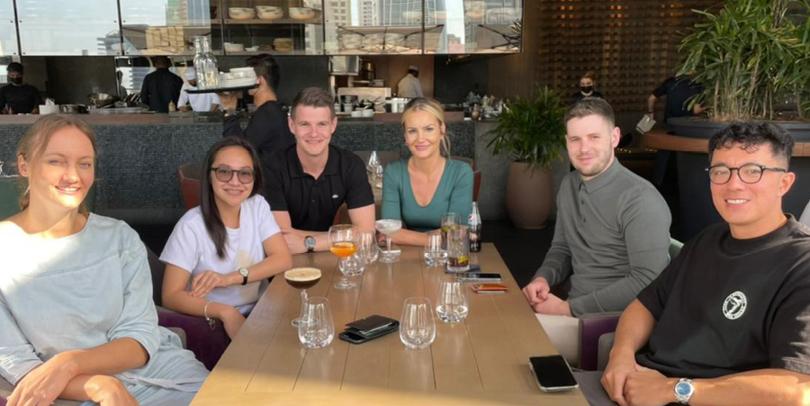 A gathering of individuals seated around a table, each has a beverage. It's a casual restaurant setting.