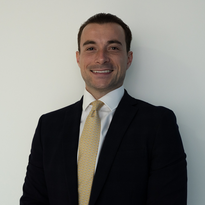 A professional young man wears a formal black suit and a gold tie, displaying a pleasant smile and looking directly at the camera. The photograph is a headshot.