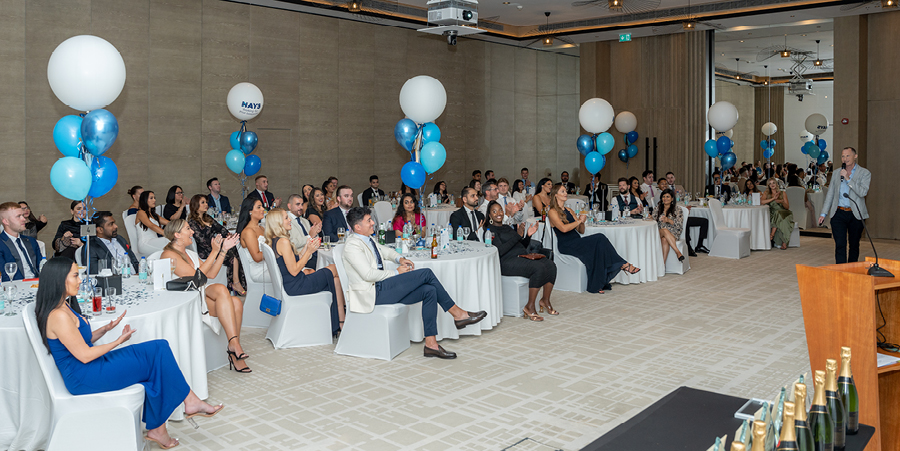 A large gathering of people in a conference room sit around round tables adorned with blue and white balloons. One man stands at the front of the room and speaks into a microphone.